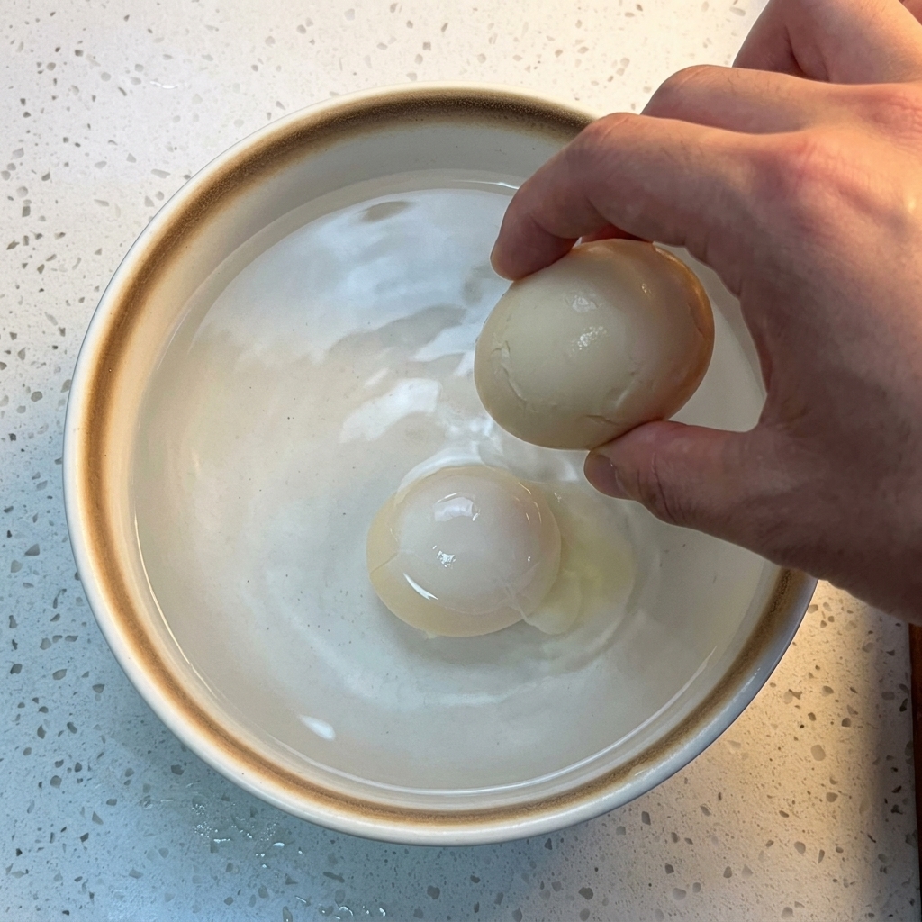 A cooked egg being submerged into a white ceramic bowl filled with cold water and ice cubes.