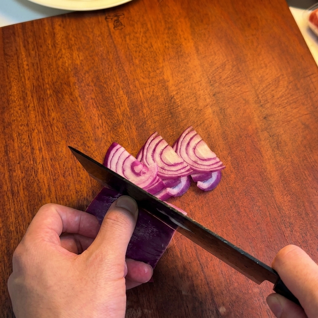 A person using a dark chefs knife to slice a purple red onion into thin wedges on a wooden board.