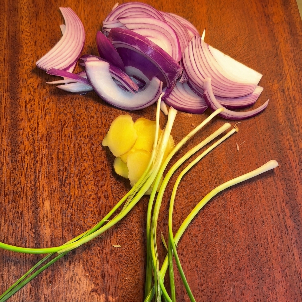 A wooden cutting board displaying sliced red onions, ginger rounds, and fresh green onion stalks.