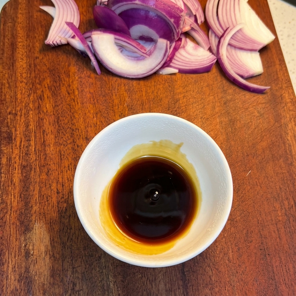A small white bowl on a wooden board containing a dark liquid sauce base, with sliced red onions visible in the background.