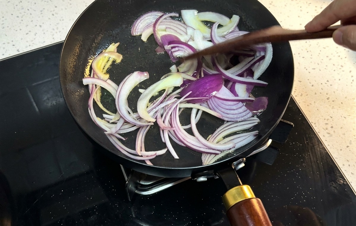 Red onion slices being stirred with a wooden spatula in a black frying pan.