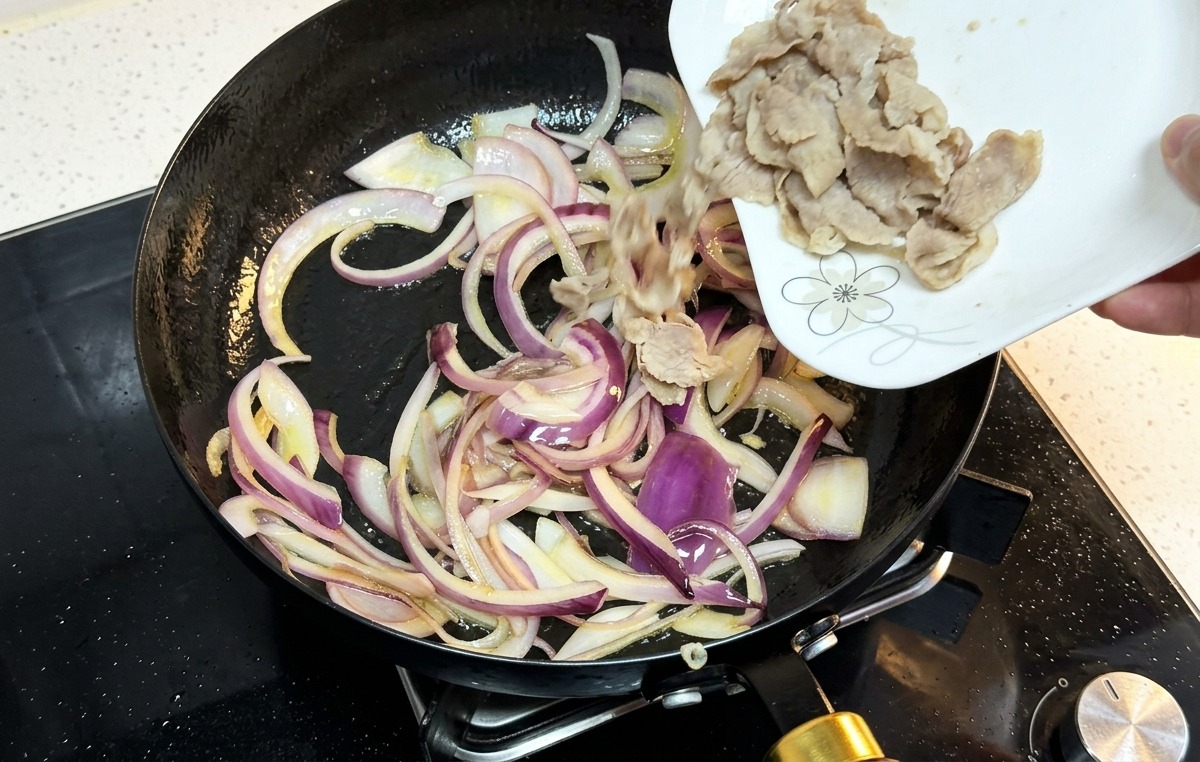 Cooked beef slices being poured from a white plate into a pan containing sautéed red onions.