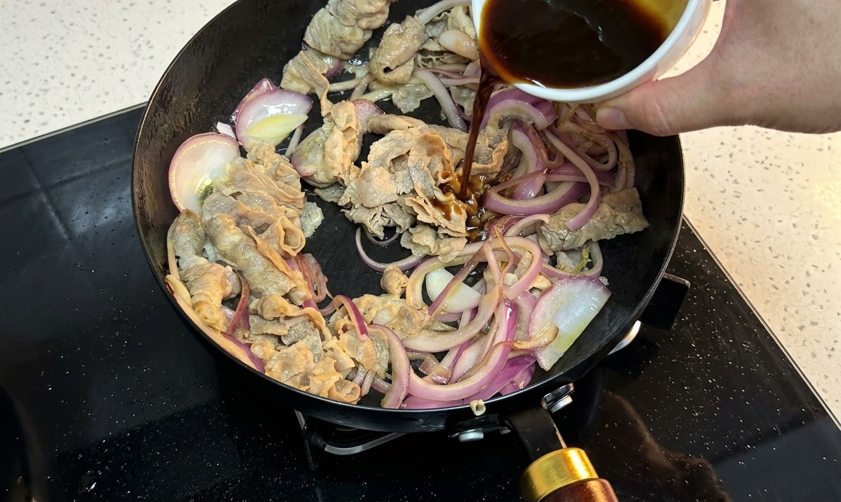Dark savory sauce being poured from a white ceramic bowl into a pan containing sliced beef and red onions.