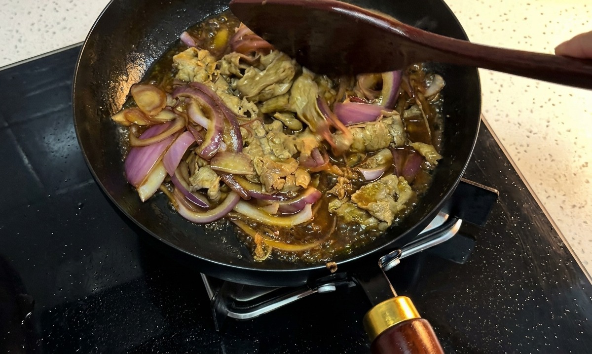 Sliced beef and red onions simmering in a thick, bubbling dark sauce inside a black pan, being stirred with a wooden spatula.