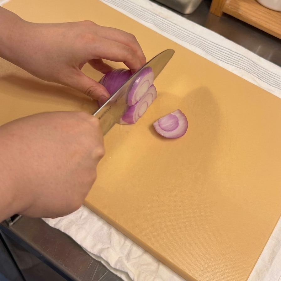 A close-up view of hands slicing a raw purple onion into thin strips on a tan cutting board.