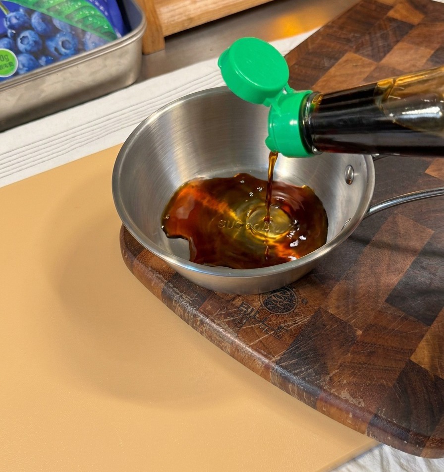 Dark soy sauce being poured from a bottle with a green cap into a small stainless steel bowl.