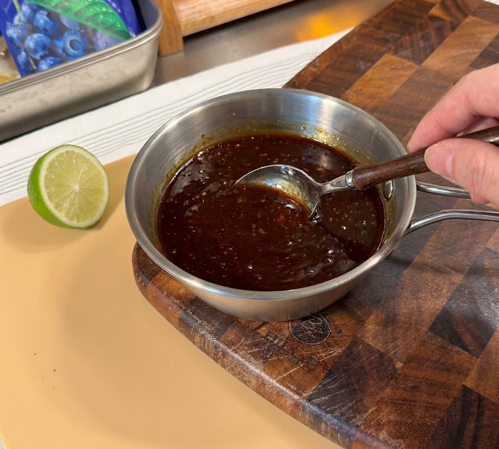A hand using a spoon to stir a thick, dark brown teriyaki sauce mixture in a small metal pot.