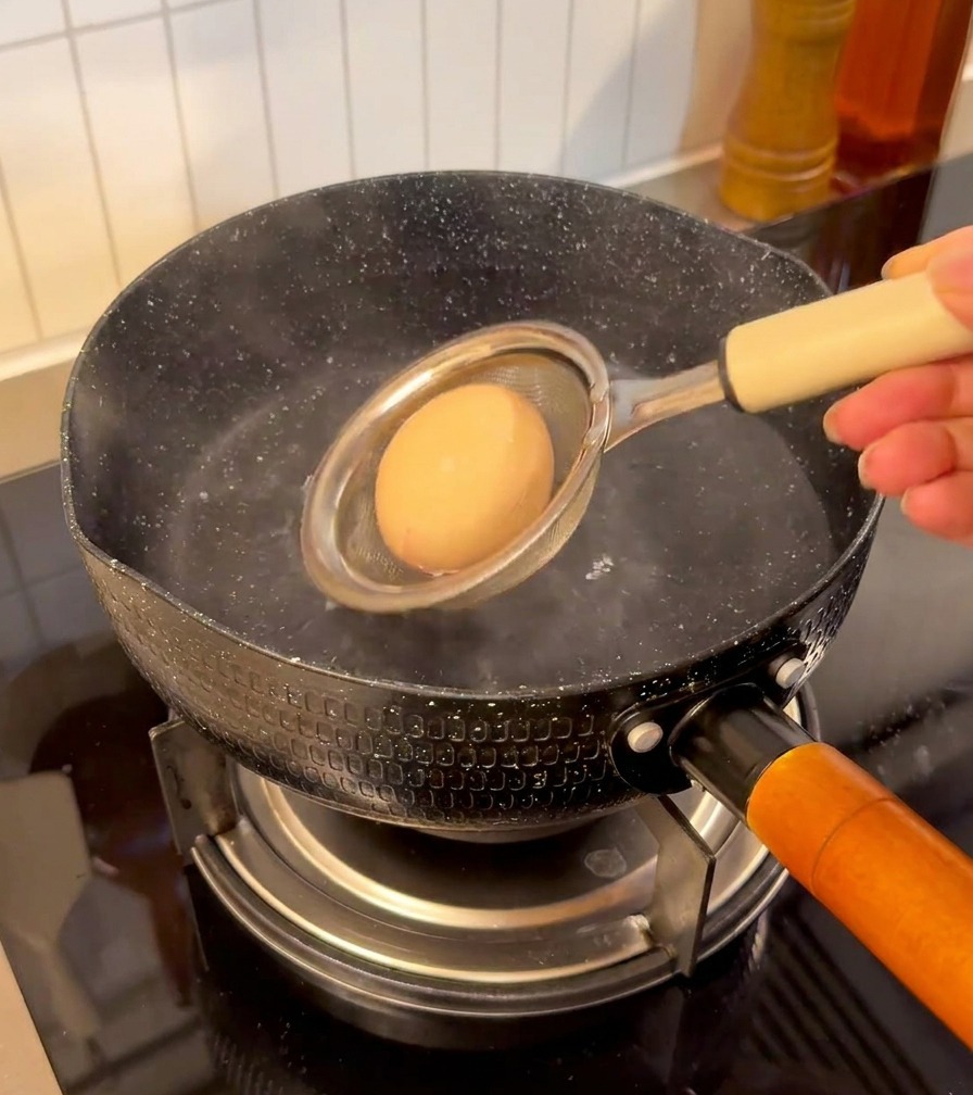 An egg held in a small mesh strainer being lowered into a pot of steaming water on a stovetop.