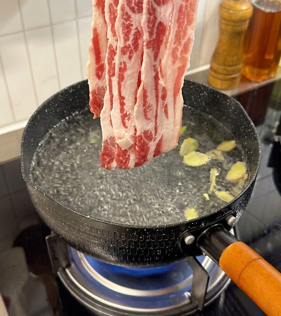 Thinly sliced raw beef being added to a pot of boiling water containing ginger slices.