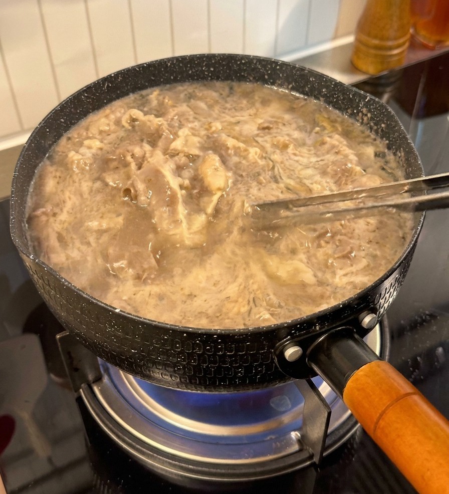 Thinly sliced beef being lifted out of a pot of boiling water with tongs appearing pale and par-cooked.