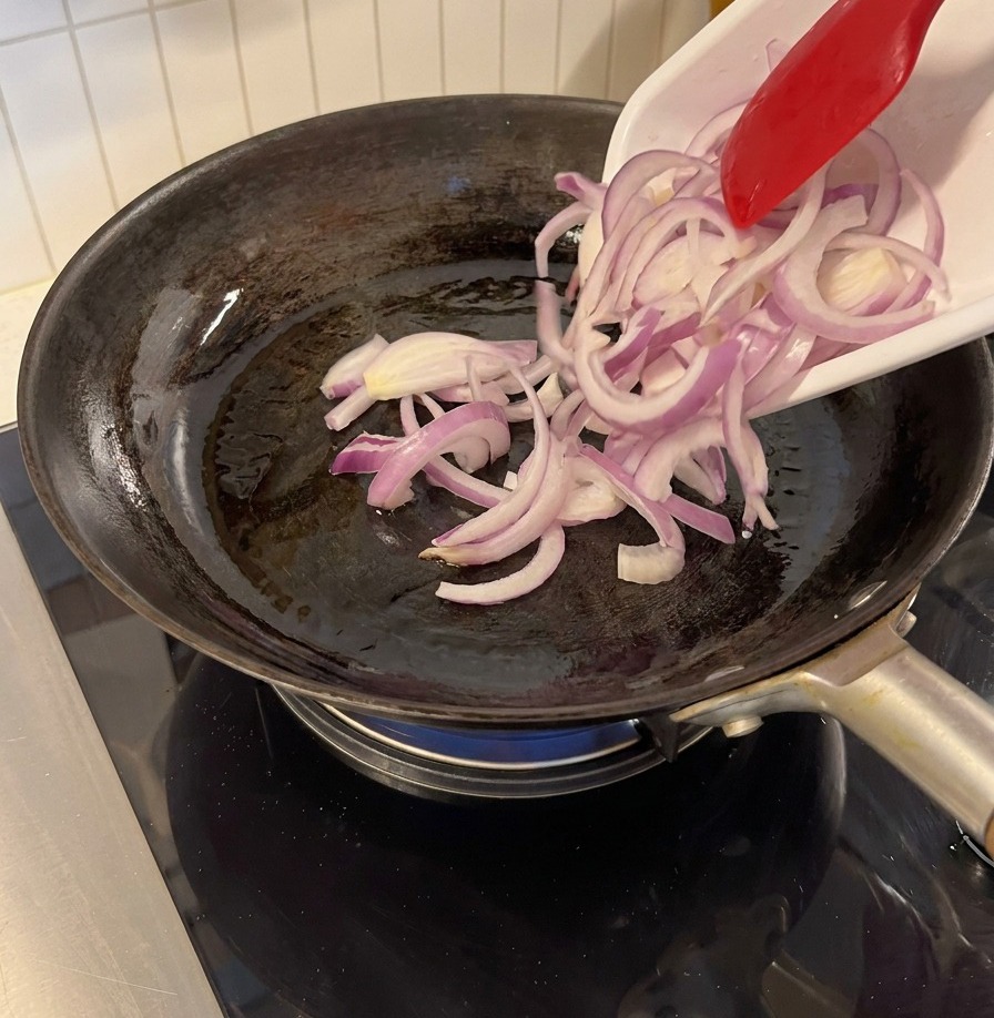 Sliced red onions being added to a hot black frying pan using a red silicone spatula.