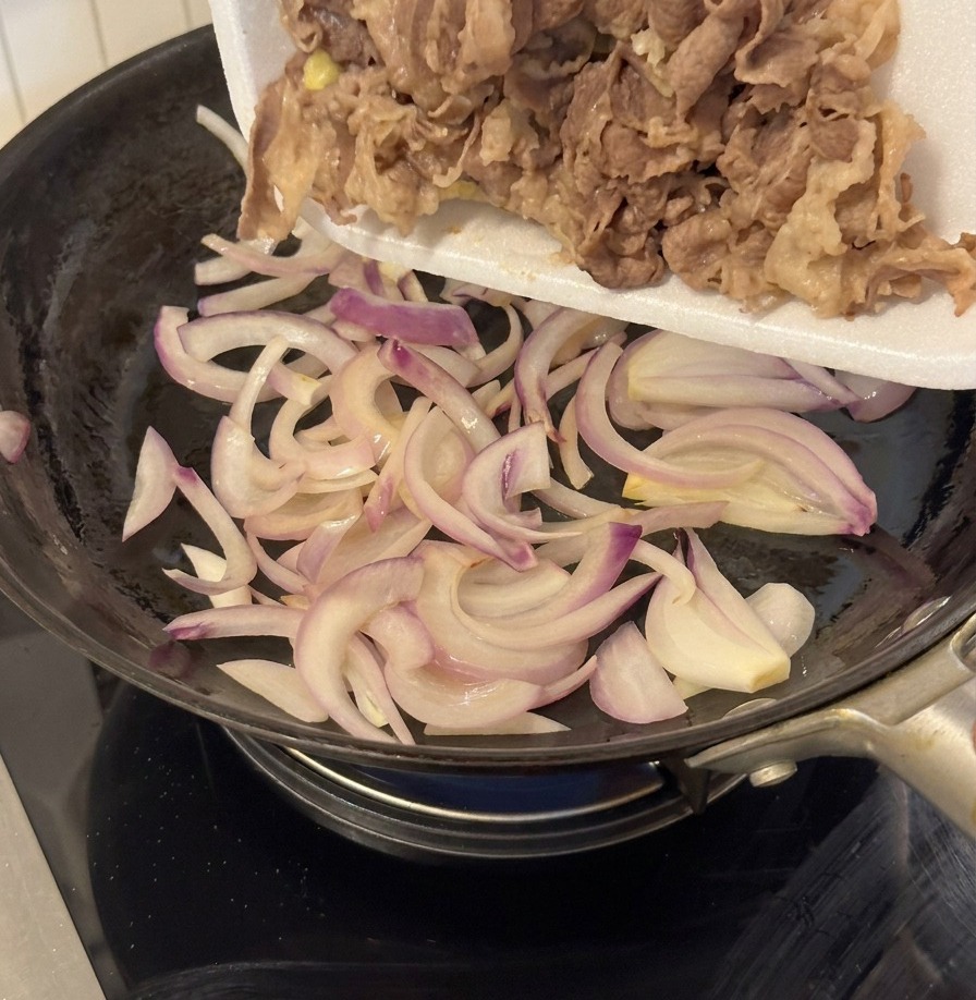 Blanched beef being poured from a white tray into a pan containing sautéed onions.