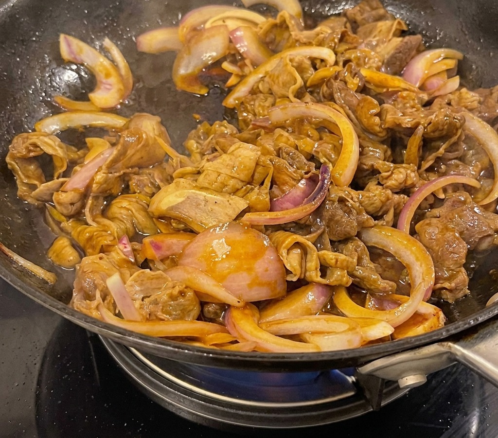 Teriyaki beef and onions simmering in a thick, dark brown glossy sauce inside a black wok.