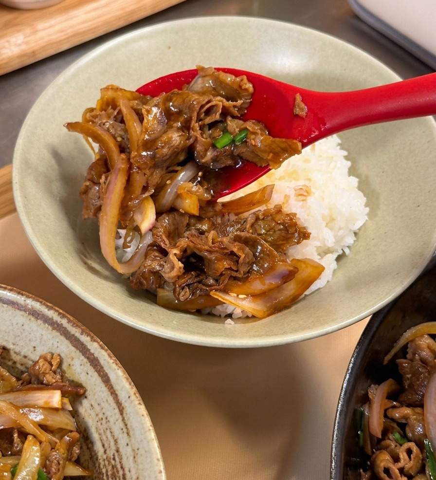 A red spoon placing a serving of glossy teriyaki beef and onions onto a bed of white rice in a ceramic bowl.
