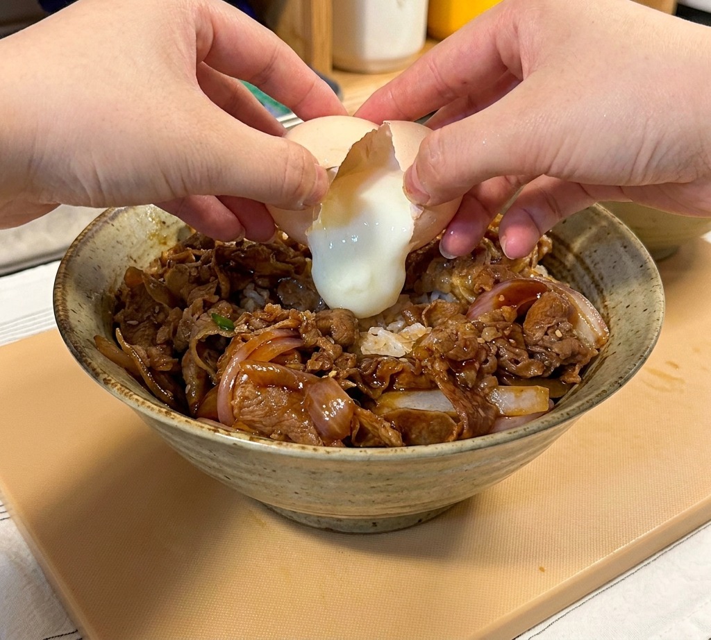 A close-up shot of two hands cracking a soft-cooked onsen egg over a bowl of sliced teriyaki beef and onions served on a bed of white rice.