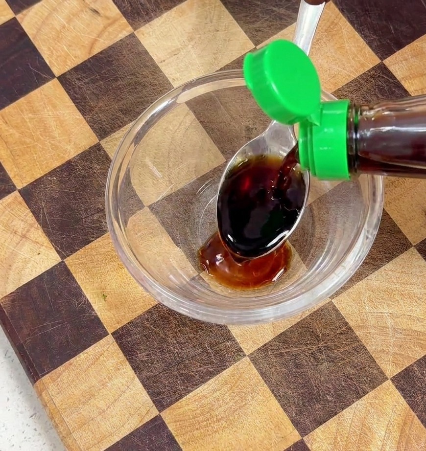 Dark liquid seasoning being poured from a bottle into a glass bowl containing soy sauce.