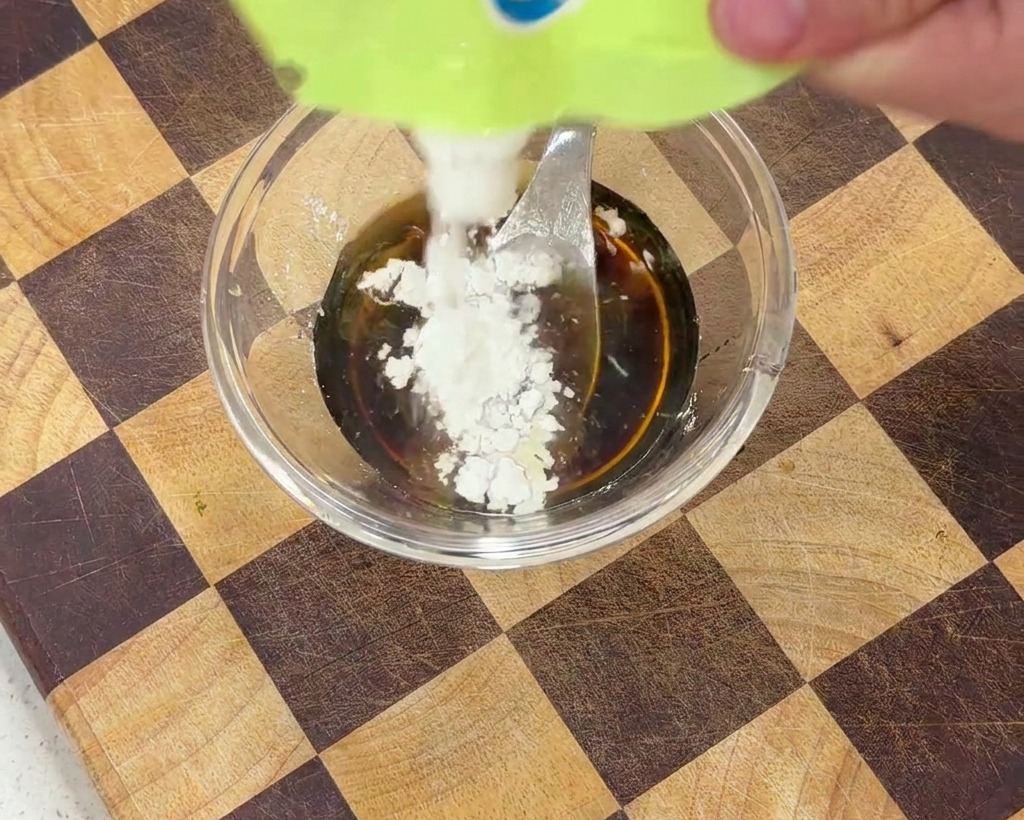 A small amount of white cornstarch being added to a dark liquid sauce in a clear bowl.