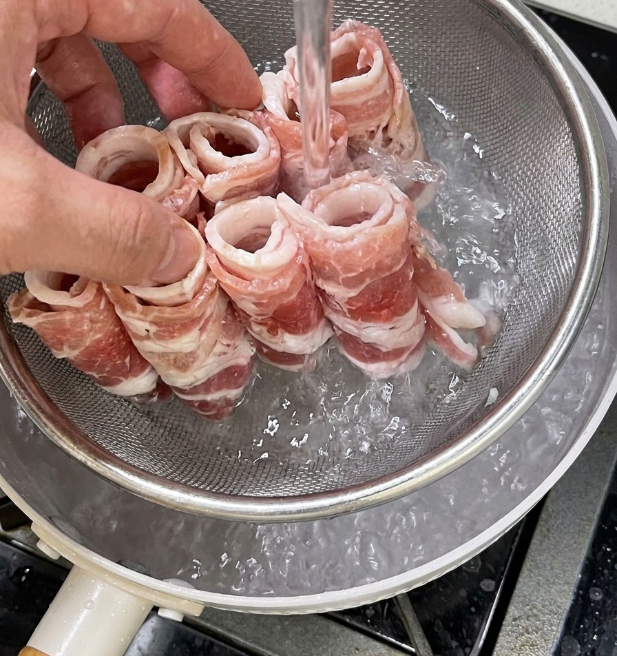 A hand holding a metal mesh strainer filled with raw beef rolls being lowered into a pot of boiling water to be blanched.