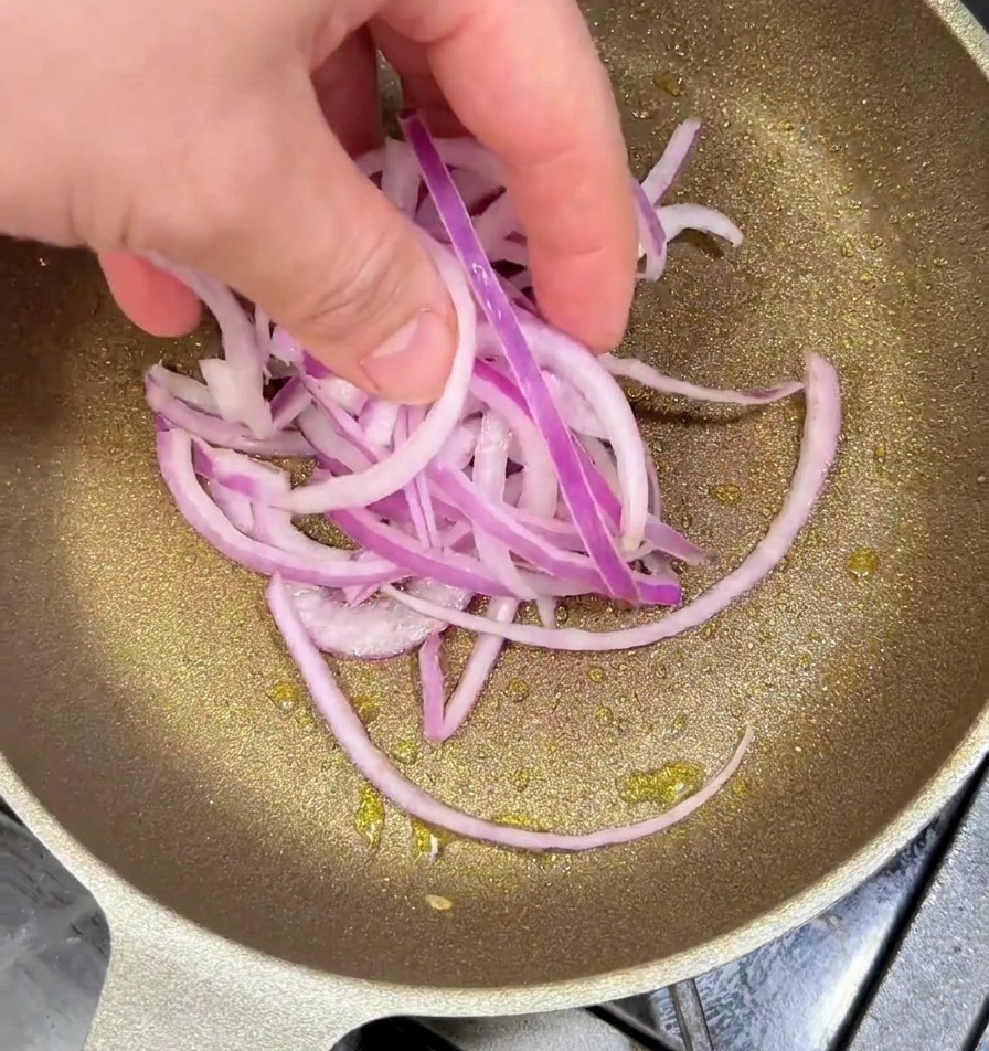 Sliced purple onions being added by hand to a heated frying pan with oil to be sautéed until charred.
