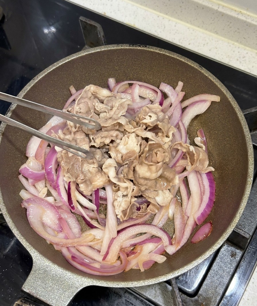 Blanched beef slices being added to a pan of sautéed purple onions using metal tongs.