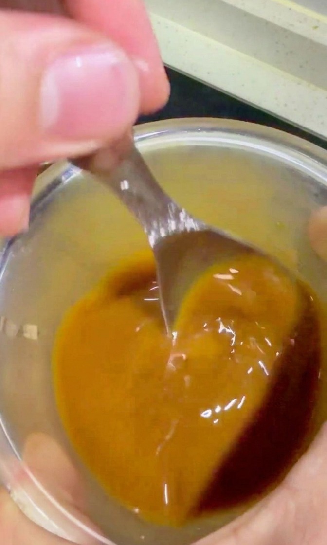 A hand using a spoon to prepare and pour a golden-brown teriyaki sauce from a glass bowl into a pan of beef and onions.