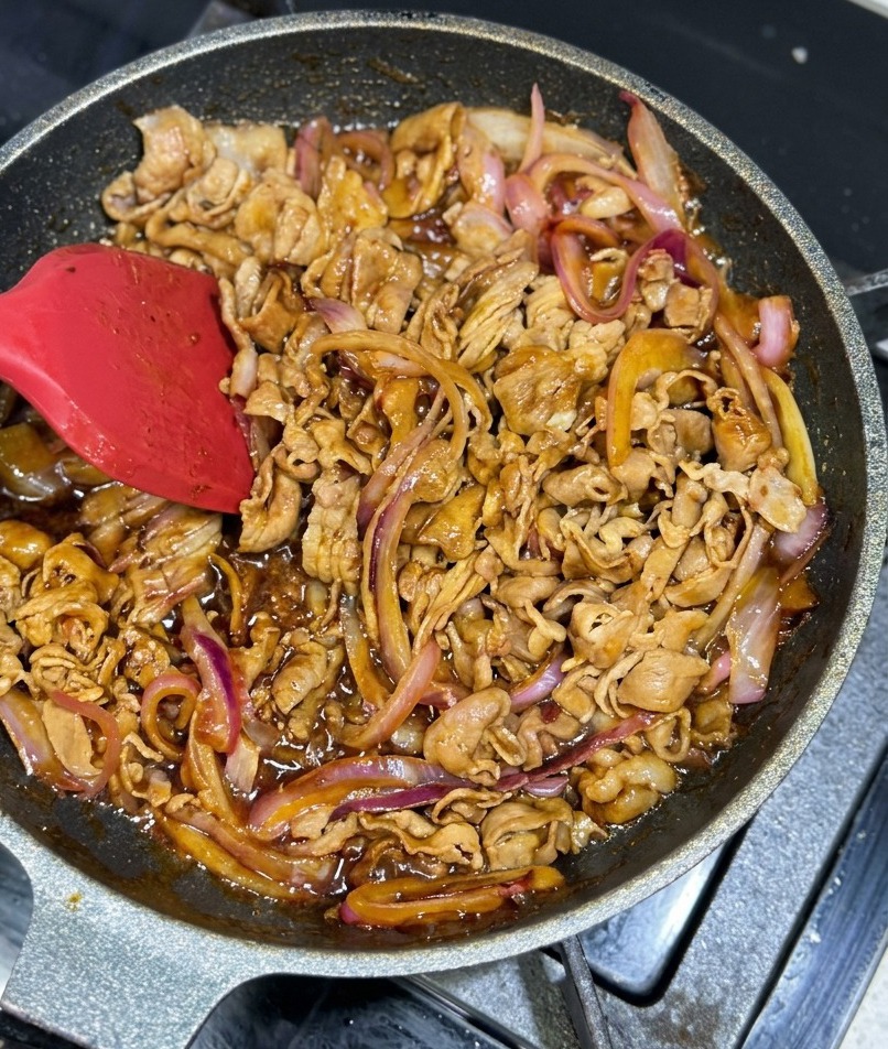 Thinly sliced beef and red onion strips being tossed in a pan with a thick, shimmering brown glaze.