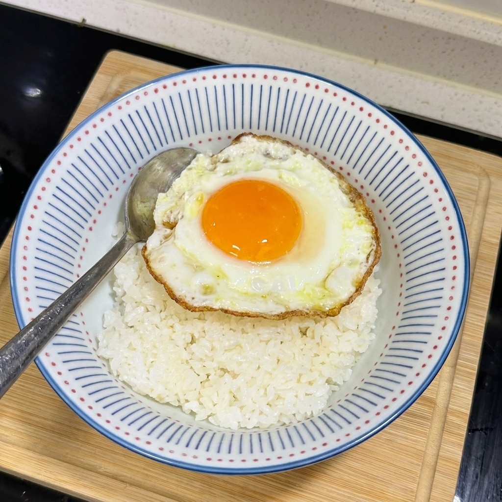 A top-down view of a white ceramic bowl filled with fluffy white rice, topped with a single fried egg featuring a vibrant orange runny yolk and slightly browned, crispy edges.