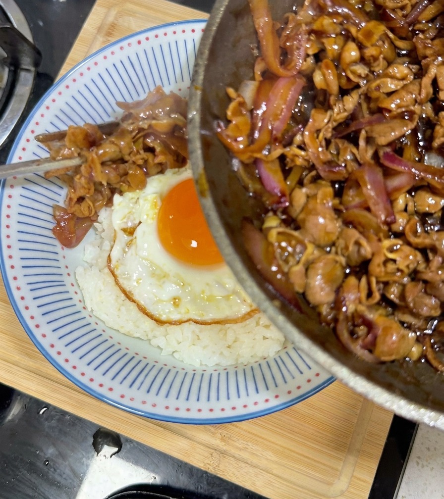 Glazed teriyaki beef and onions being poured from a frying pan onto a bowl of white rice topped with a fried egg.