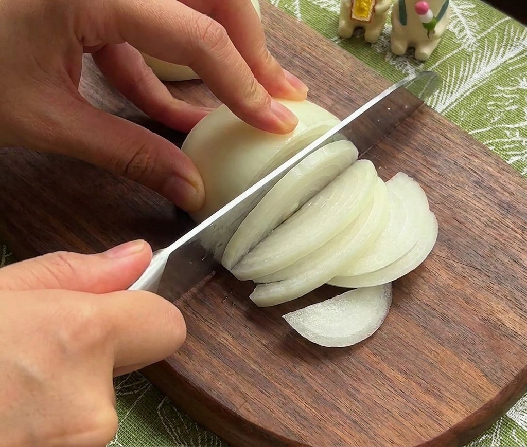 A persons hands using a chefs knife to slice a white onion into thin strips on a dark wooden cutting board.