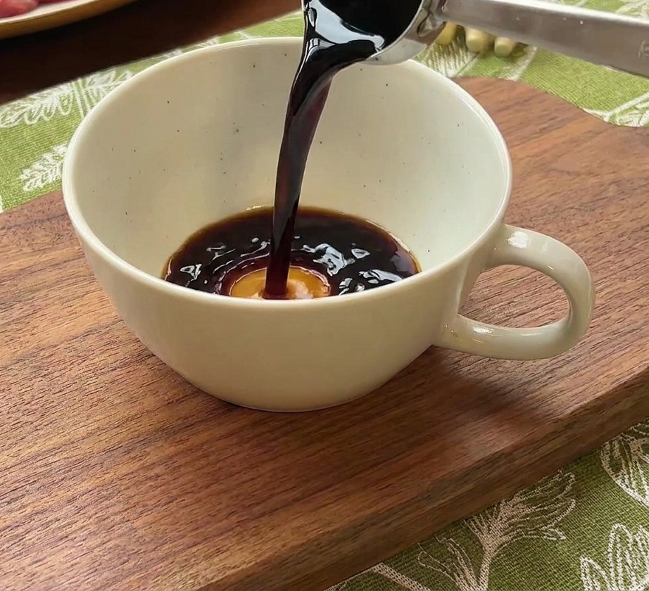 A metal spoon pouring dark soy sauce into a white ceramic bowl containing water and a dollop of red Korean chili paste.