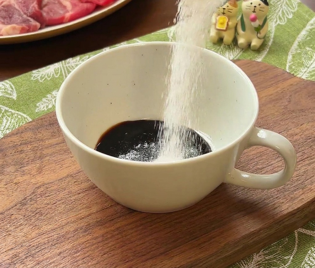 White granulated sugar being poured from a metal measuring scoop into a white bowl already containing a dark liquid sauce base.