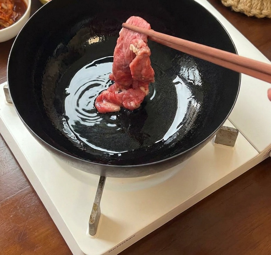Thin slices of fresh raw beef being placed into a hot, oiled iron pan using chopsticks.
