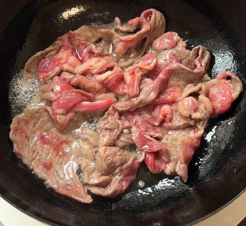 Sizzling browned beef slices being moved from a dark pan to a white ceramic plate.