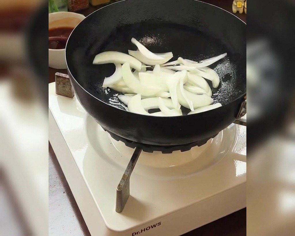 Sliced white onions being added to a pan with a bowl of red kimchi visible in the background.