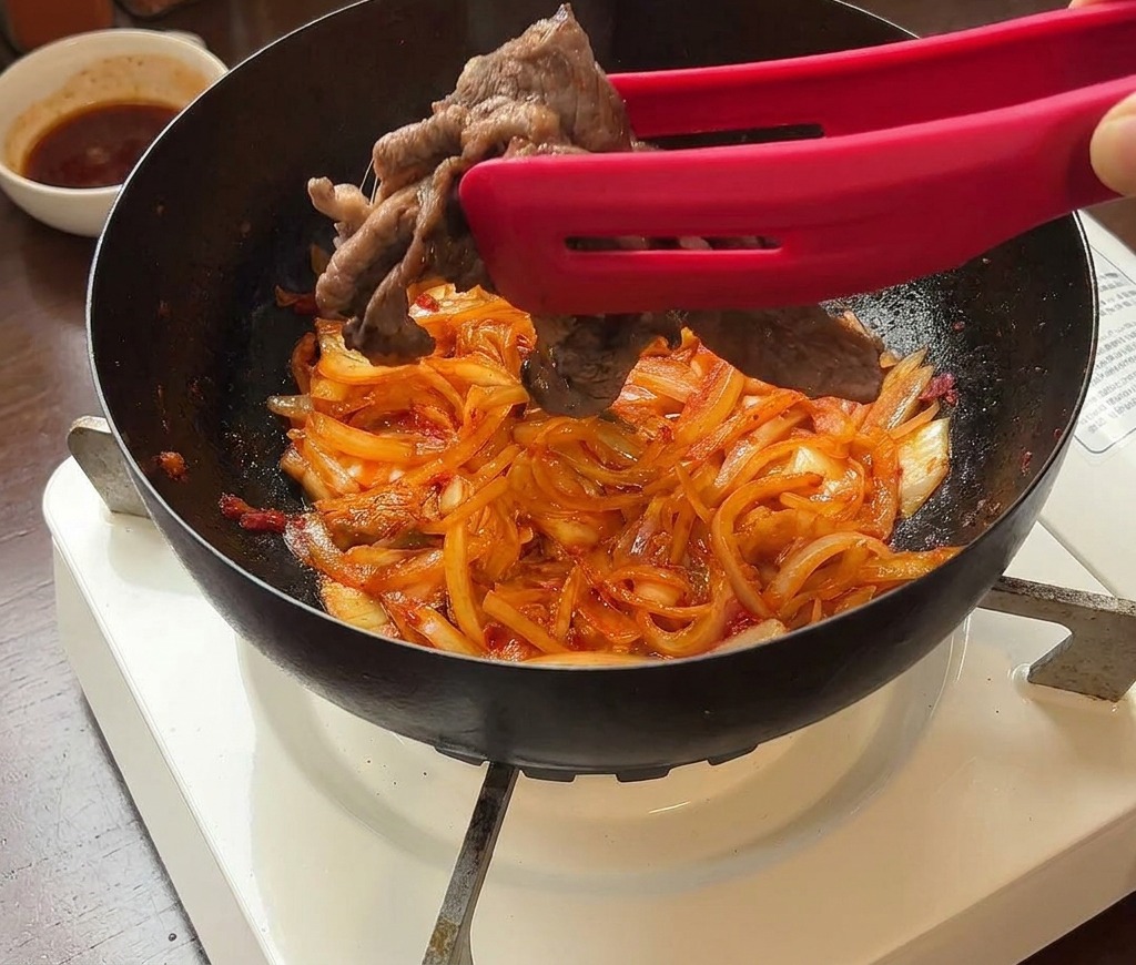 Seared beef slices being added back to a pan containing stir-fried kimchi and onions using red tongs.