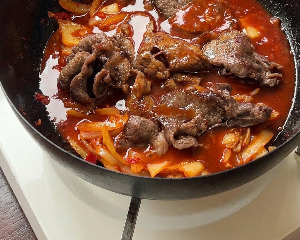 The beef and kimchi mixture simmering in a thick, vibrant red sauce within a black iron pan.