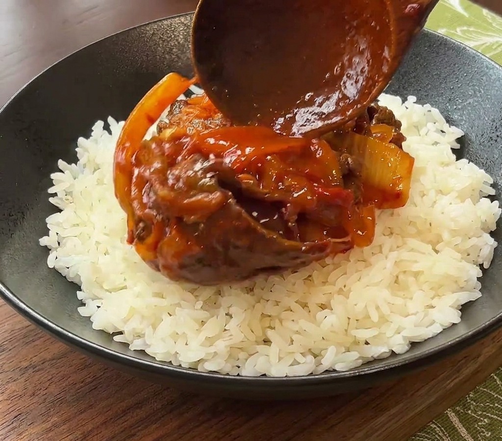 A wooden spoon placing a serving of cooked beef and kimchi in red sauce onto a bed of white rice in a black ceramic bowl.