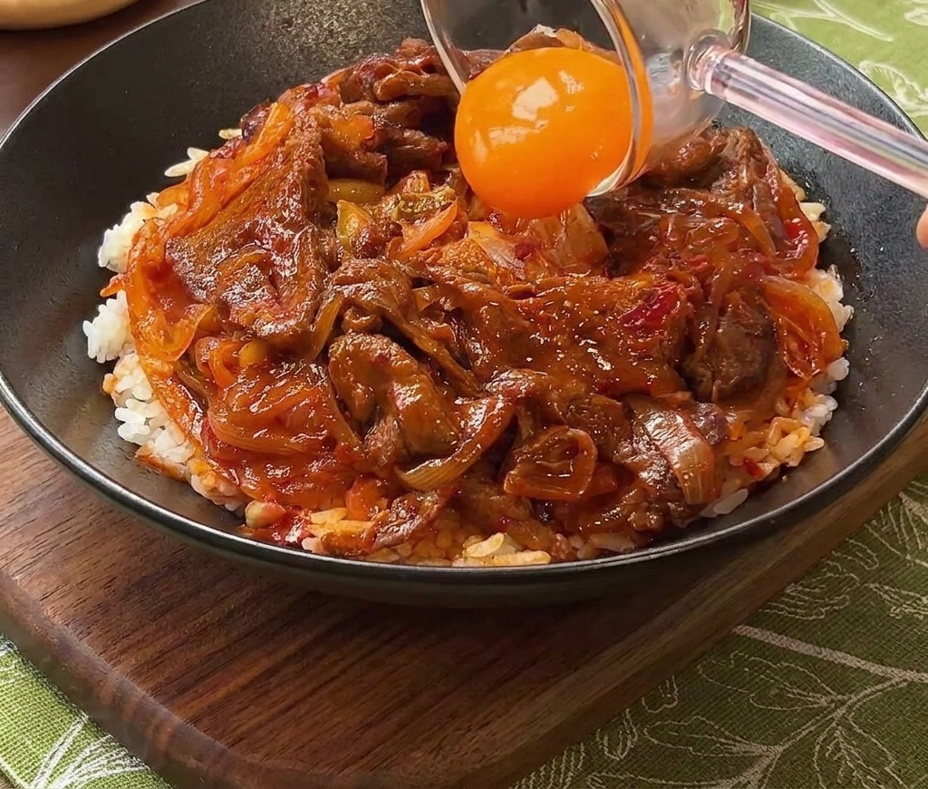 A bright orange egg yolk being placed onto a bowl of beef and rice using a glass separator.