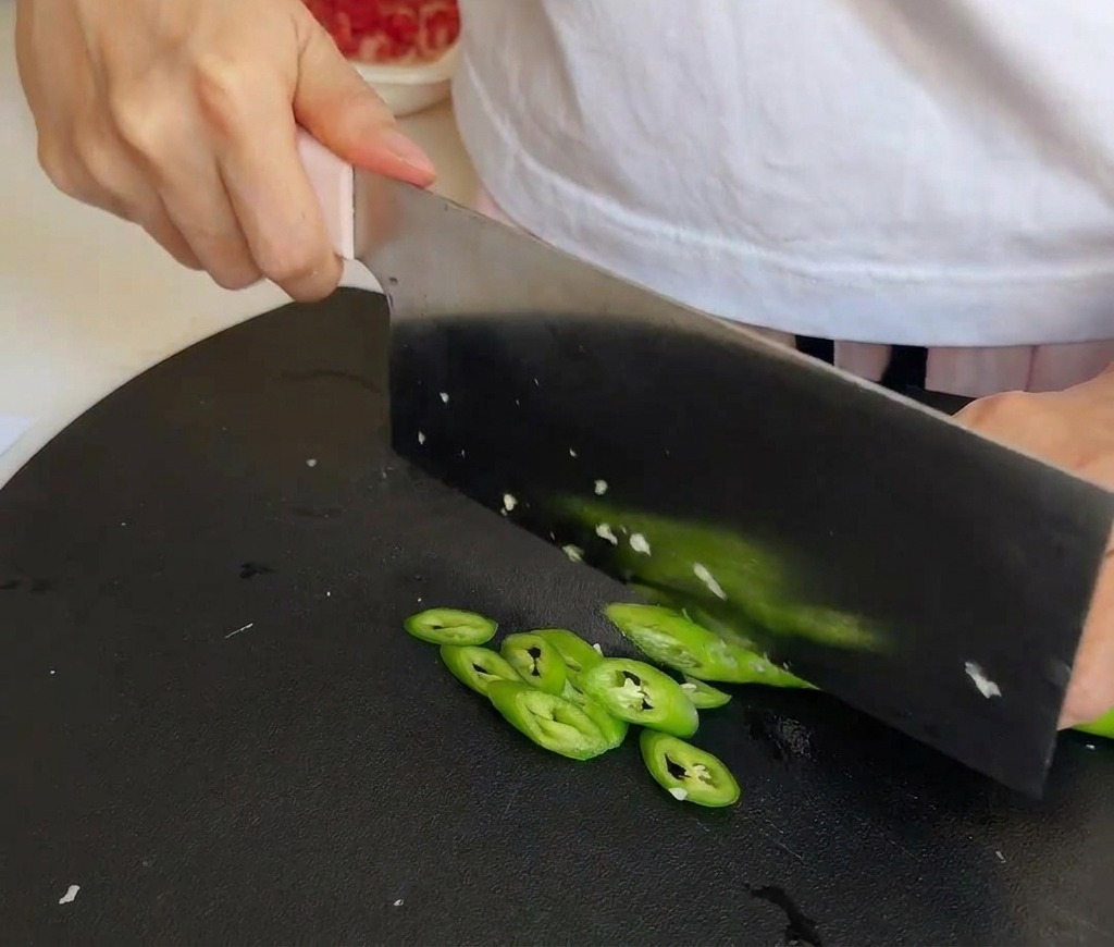 A kitchen cleaver slicing through bright green chili peppers on a textured black cutting board.