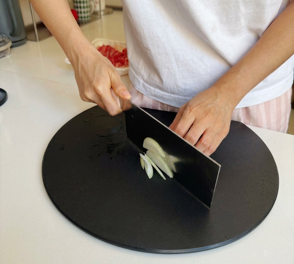 A persons hands using a large kitchen cleaver to slice a white onion into thin strips on a black circular cutting board.