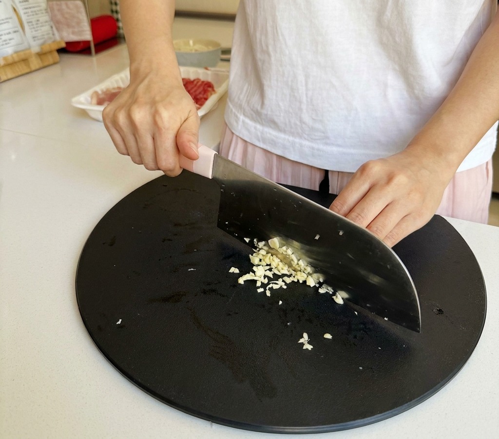 A close-up of a persons hands mincing garlic cloves into fine bits on a black cutting board using a chefs knife.