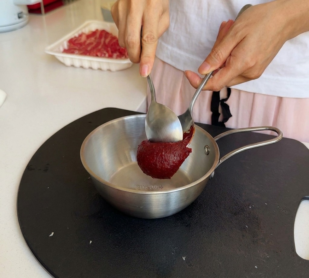 A person using two metal spoons to add a large scoop of deep red Korean chili paste into a stainless steel bowl.