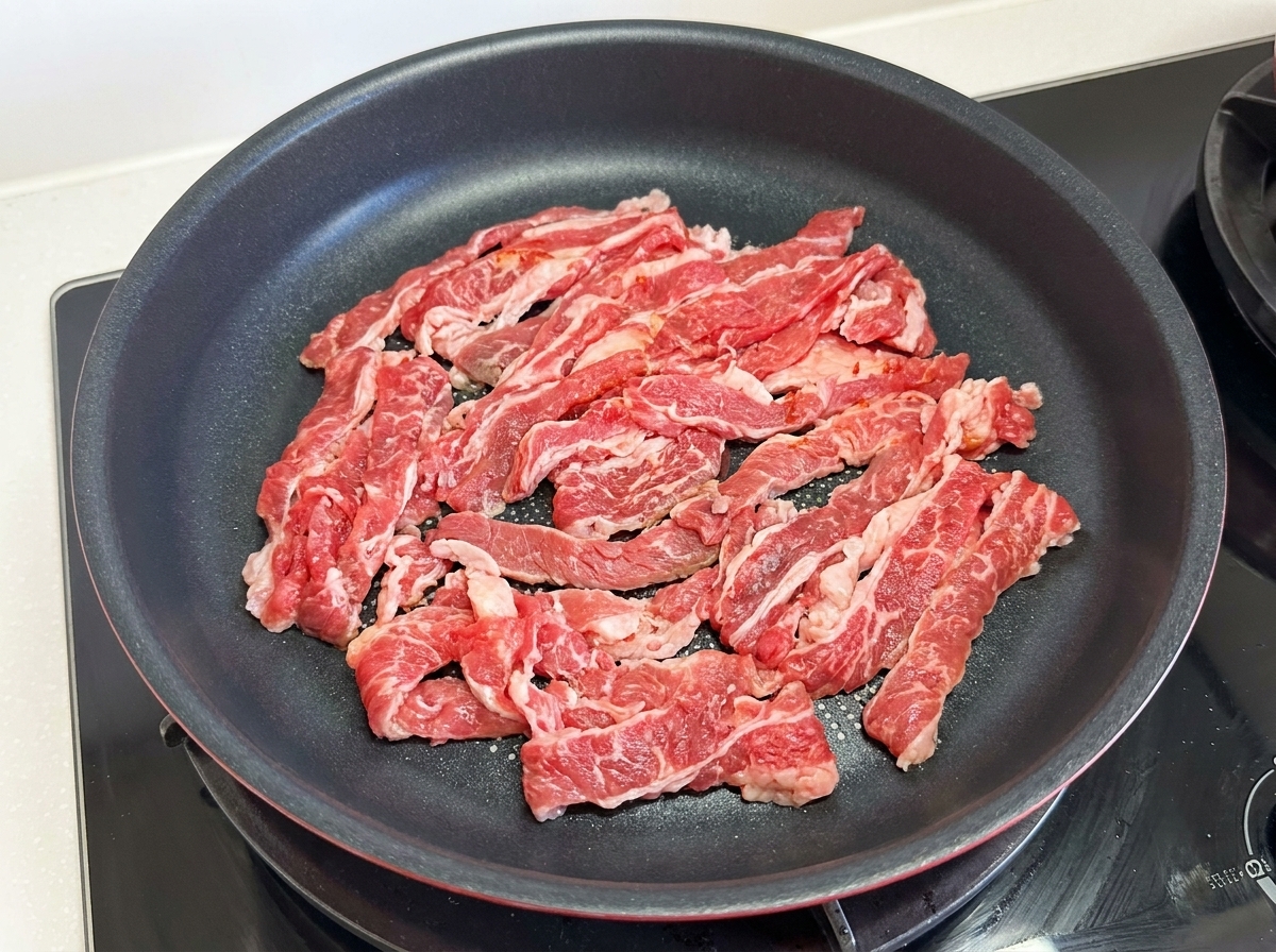 Thinly sliced, raw fat beef pieces spread out across the surface of a black non-stick frying pan on a stovetop.
