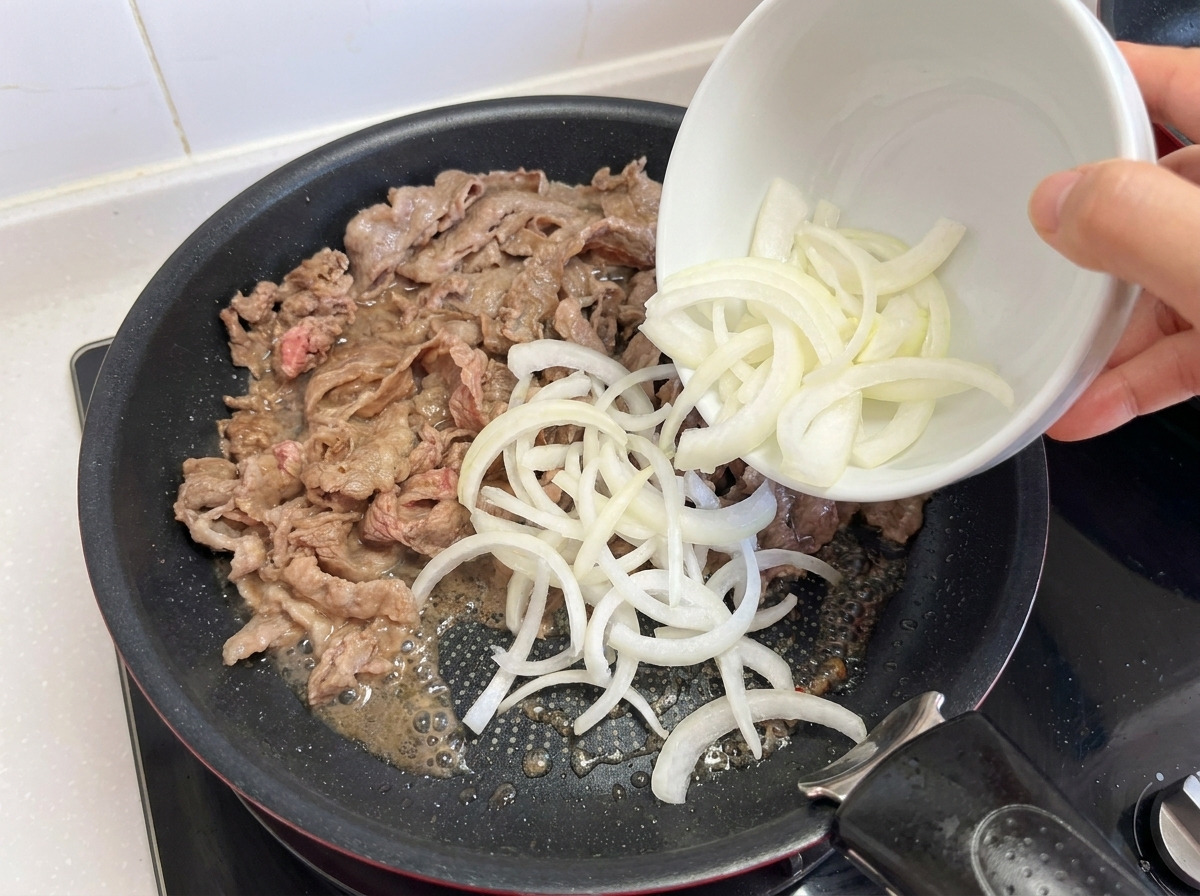 A hand pouring a bowl of sliced white onions into a frying pan that contains partially cooked strips of beef.