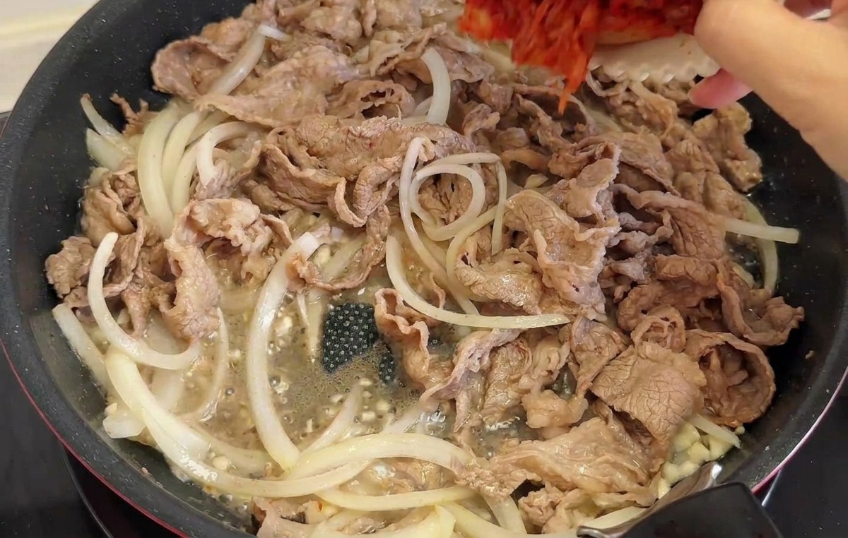A hand pouring a white bowl filled with red chopped kimchi and green pepper slices into a skillet containing stir-fried beef and onions.