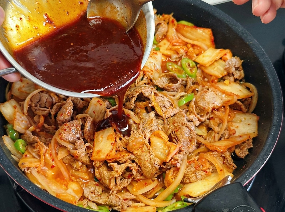 A person pouring a dark red sauce from a small metal bowl into a frying pan containing beef slices, onions, kimchi, and green chilies.
