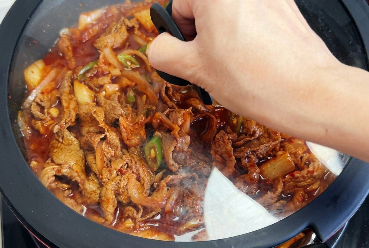 A hand placing a black-rimmed glass lid over a frying pan filled with simmering kimchi beef and peppers in a red sauce.