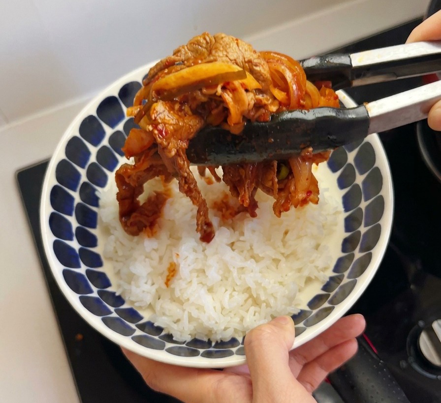 Black tongs holding a serving of cooked beef and red kimchi, placing it onto a bowl of white rice.