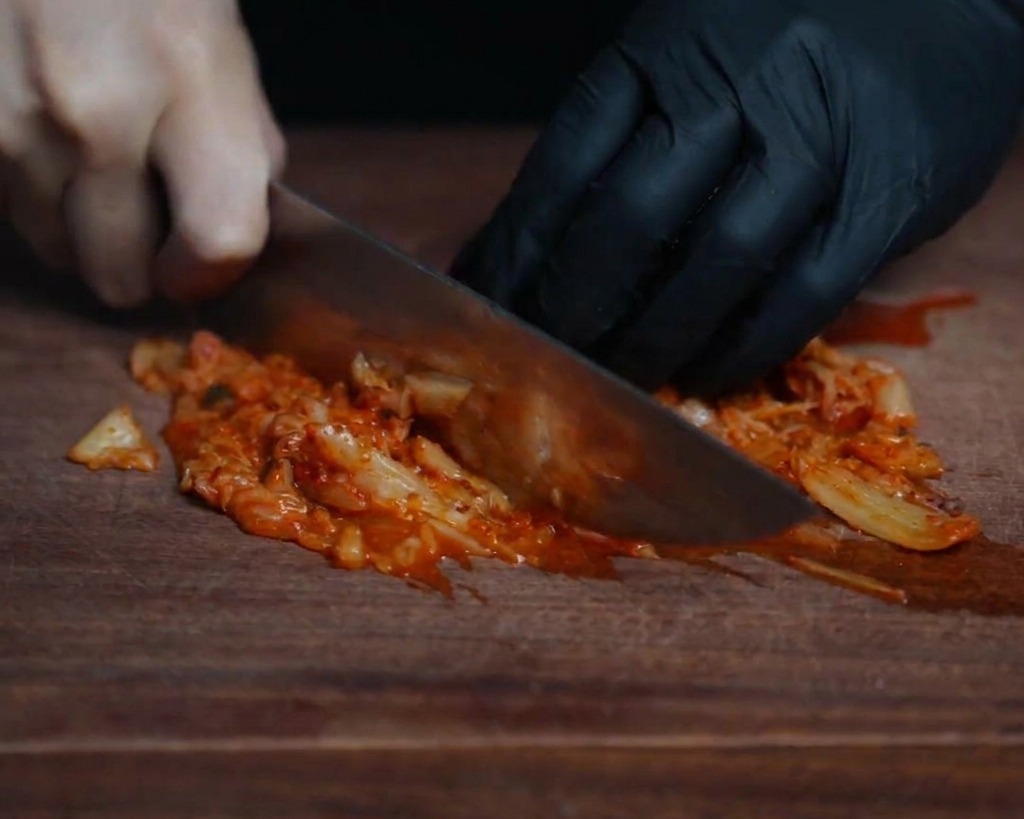 Close-up of a chef finely chopping bright red fermented kimchi on a dark wooden board with a sharp knife.