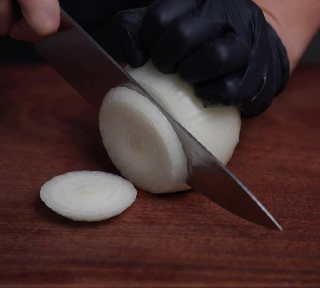 A white onion being sliced into thin, translucent rings on a wooden surface using a professional chefs knife.